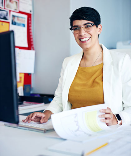 Mujer con blazer blanco y gafas sonriendo frente a un ordenador, sosteniendo unos documentos